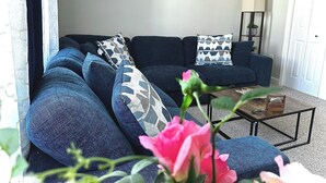 A living room with a dark blue sofa, patterned cushions, a coffee table, and a painting on the wall.