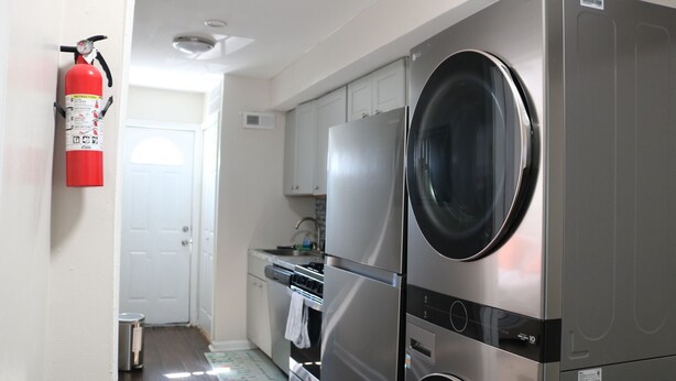 A compact kitchen with a stainless steel washer and dryer, a white door, and a fire extinguisher mounted on the wall.