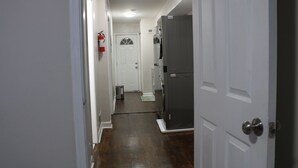 A narrow hallway with a white door at the end, a fire extinguisher on the wall, and a large gray refrigerator.