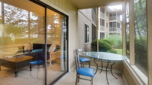 A glass-enclosed balcony with a table and chairs, a wooden bench, and a view of a multi-story building and greenery. - 5 Mi to Cherry Grove Beach: Dog-Friendly Condo! (Little River)
