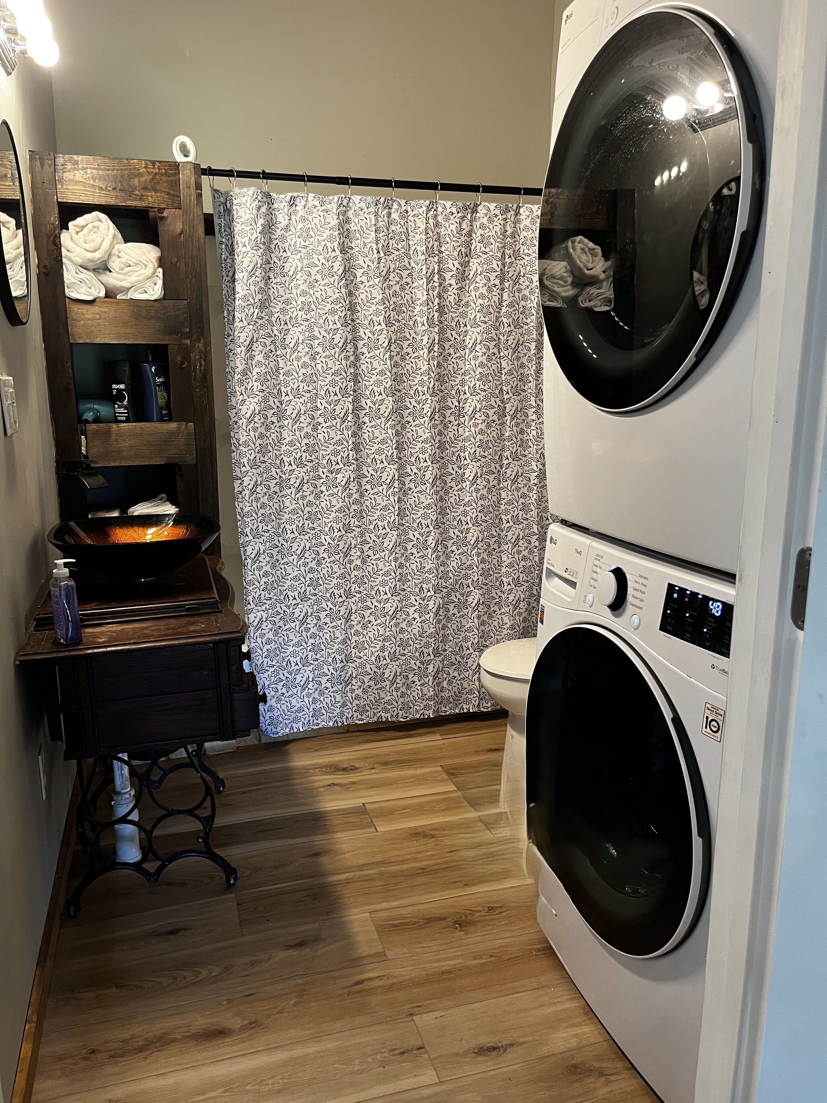 A laundry room with a double washer and dryer, a wooden shelf with towels, and a patterned shower curtain.