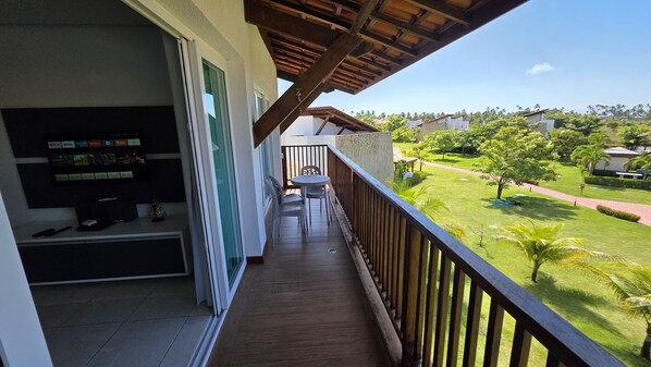 A balcony with a TV, a table, and chairs, overlooking a well-maintained lawn and buildings in the distance. - Club Meridional Carneiros 01PS byLP (Tamandaré)