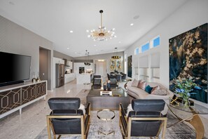 A modern living room with a large TV, a dining area with a glass coffee table, and a kitchen in the background. - Modern Desert Retreat | Pool, Spa, EV Charger, Near Tennis Garden (Indian Wells)