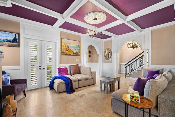 A spacious living room with a beige sofa, a patterned armchair, and a coffee table. There is a staircase with a black railing leading to an upper floor.