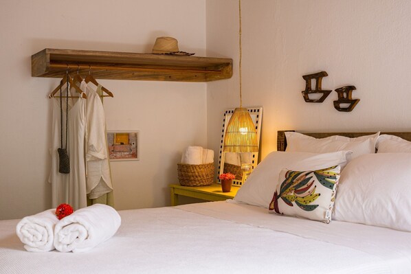 A neatly made bed with white linens, a wooden shelf above holding a hat, and a bedside table with a basket of towels and a small vase. - Casa Mangará (Cruz)