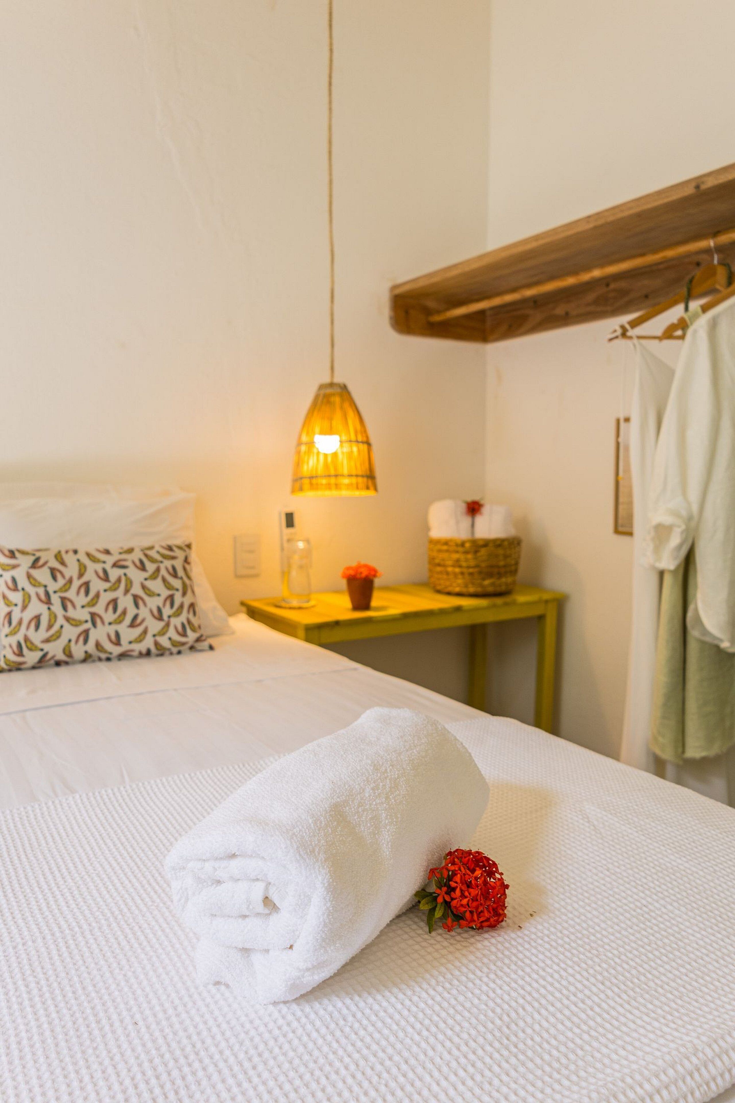 A neatly made bed with a rolled white towel and a red flower, a bedside table with a lamp, a basket, and a bottle.