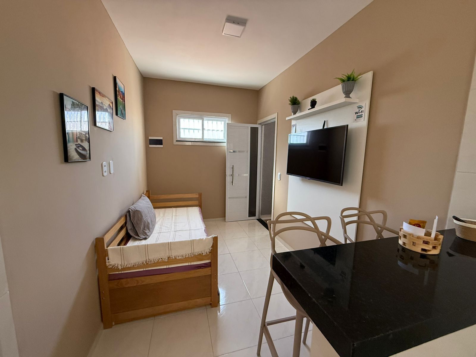 A small, well-lit room with a bed, a TV mounted on the wall, a dining table with chairs, and a window providing natural light.