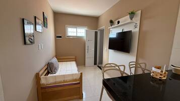 A small, well-lit room with a bed, a TV mounted on the wall, a dining table with chairs, and a window providing natural light.