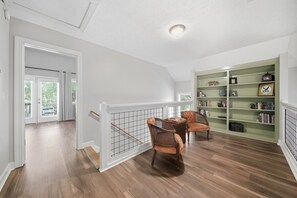 Une pièce spacieuse avec un plancher en bois, un coin repas avec deux chaises, une bibliothèque et une vue sur l’extérieur, visible à travers une porte.