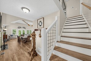 A spacious living area with a dining table, a staircase with wooden steps, and a view of the outdoors through large windows.