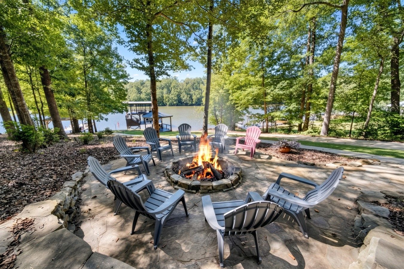 An outdoor seating area with Adirondack chairs and a fire pit, surrounded by trees and a view of a lake.