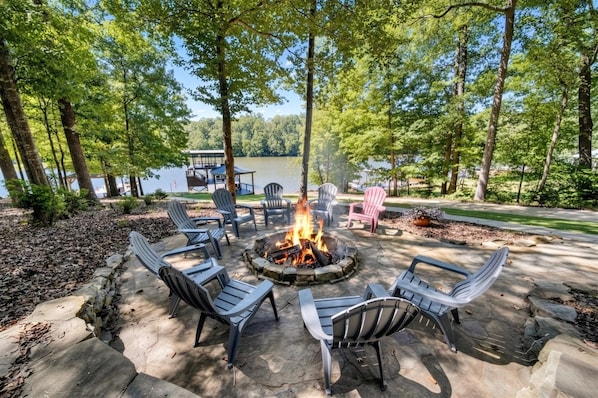 An outdoor seating area with Adirondack chairs and a fire pit, surrounded by trees and a view of a lake.