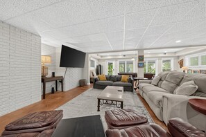 Un salon spacieux avec un mur en briques blanches, un téléviseur à écran plat fixé au mur, un canapé gris, un canapé beige et une table basse.