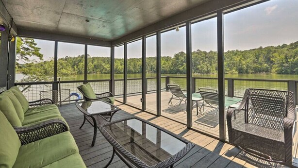 A covered outdoor deck with wicker furniture and a glass table, overlooking a lake with trees in the background.