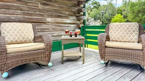 Uma área de estar ao ar livre com móveis de vime, incluindo duas cadeiras e uma pequena mesa, com uma cabana de madeira ao fundo.