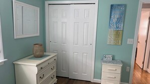 A room with teal walls, a white door, a white dresser with drawers, a wicker basket on the dresser, and a small table with a sign. - Beautifully appointed 2 bedroom with access to a private beach! (DENNIS PORT)