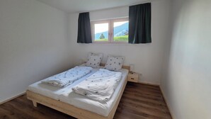 Un lit simple avec un cadre en bois, une literie blanche et deux oreillers. Il y a une fenêtre avec des rideaux, ainsi qu’une petite table de chevet avec une lampe.