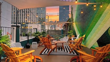 An outdoor seating area with wooden chairs and tables, string lights, and a view of city buildings.