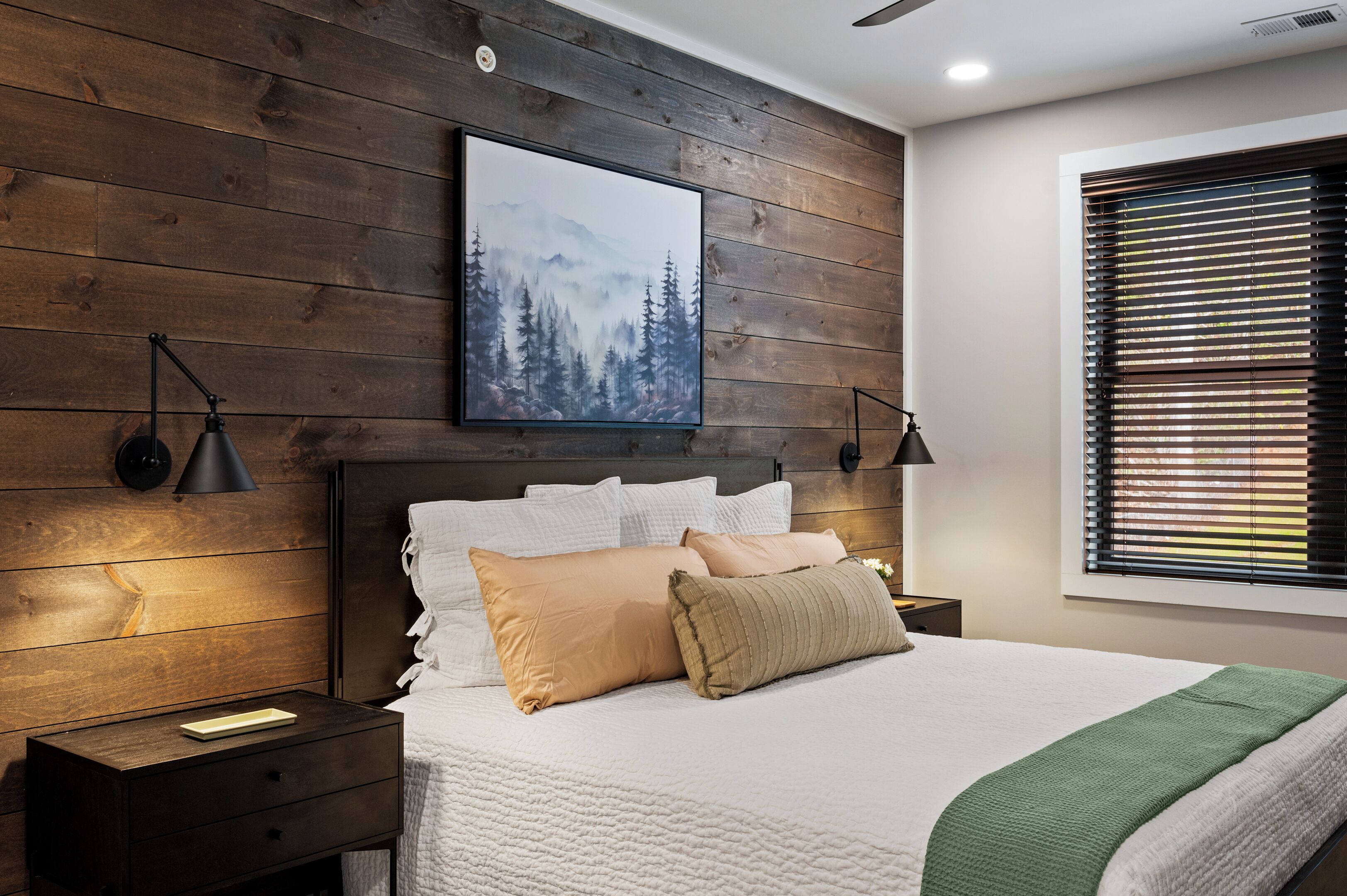Un dormitorio con cabecera de madera, una cama grande con ropa blanca, dos lámparas de noche, una mesita de noche con lámpara, un cuadro enmarcado en la pared y una ventana con persianas.