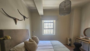 A bedroom with a bed, a bedside table, a mirror, a radiator, and a window with a view of buildings.