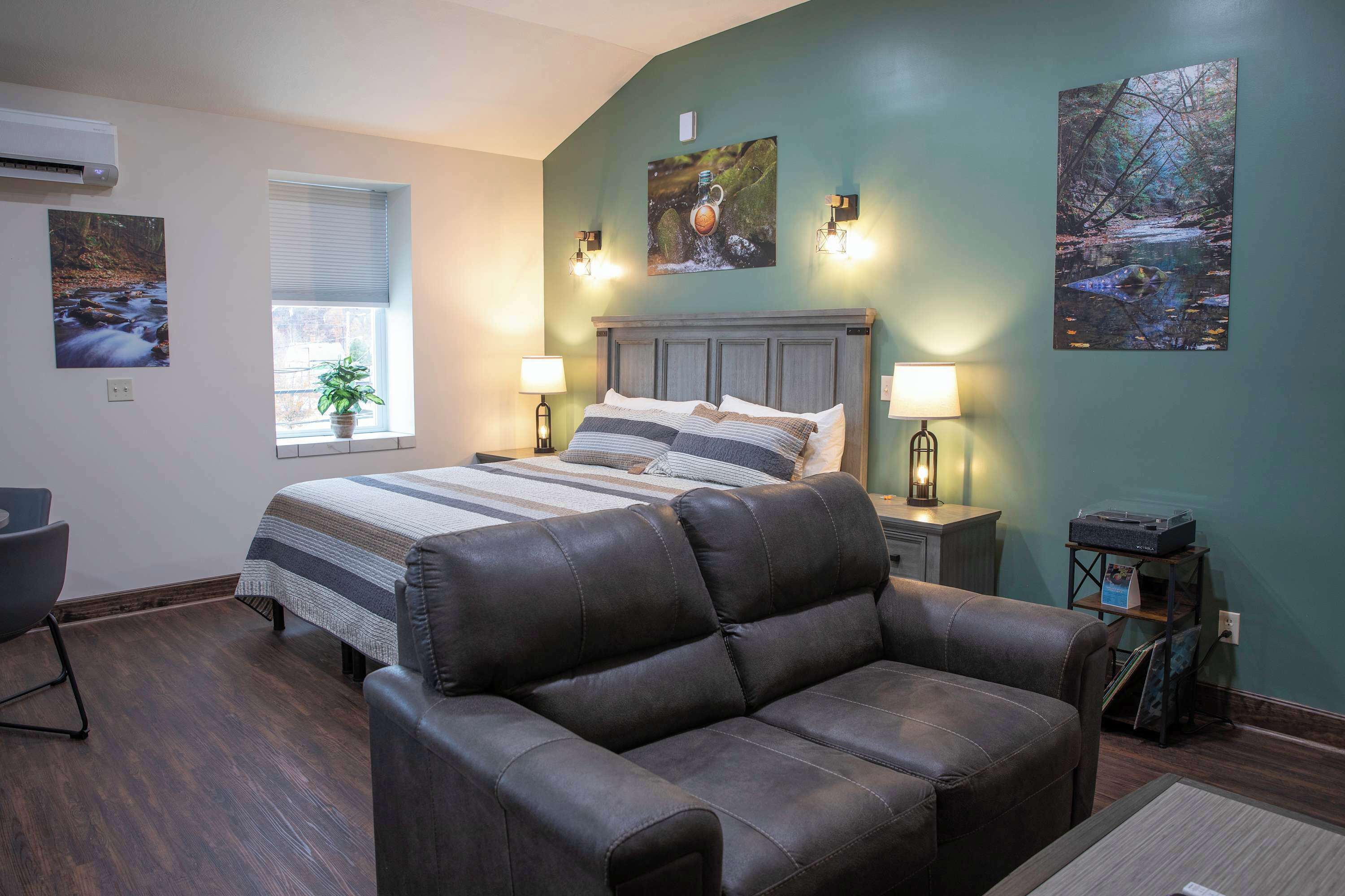 A bedroom with a bed, two bedside lamps, a window with blinds, a potted plant, and artwork on the walls.