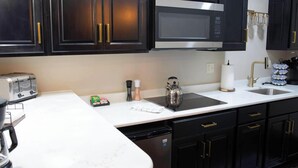 A kitchen with dark cabinets, a stainless steel microwave, a toaster, a kettle, and a sink with a gold faucet.