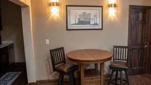 A dining area with a round wooden table and two chairs, a framed black and white photograph on the wall, and two wall-mounted light fixtures.