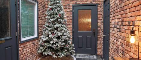 A brick-walled hallway with a frosted Christmas tree, a dark door, and a light fixture.
