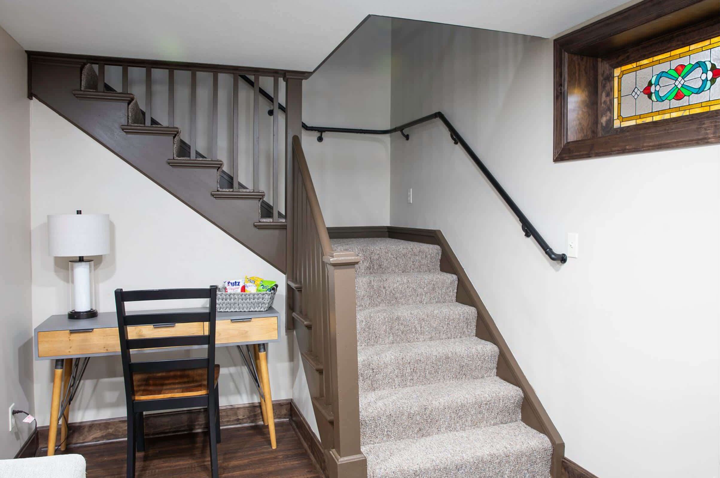 A staircase with a wooden handrail and carpeted steps, a desk with a chair, and a wall-mounted stained glass window.