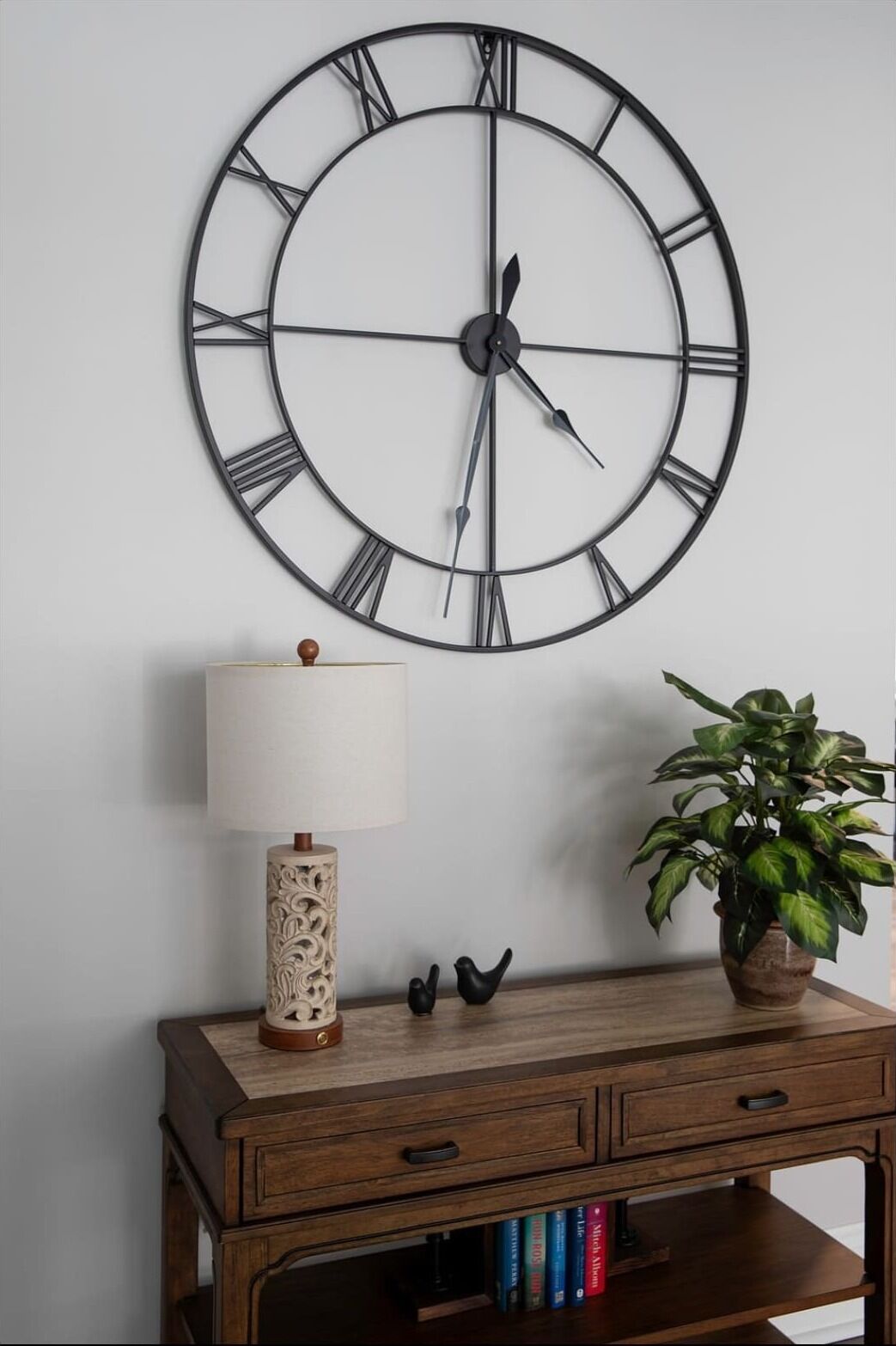 A wall-mounted clock with Roman numerals, a lamp, a potted plant, and a wooden side table with decorative items and books.