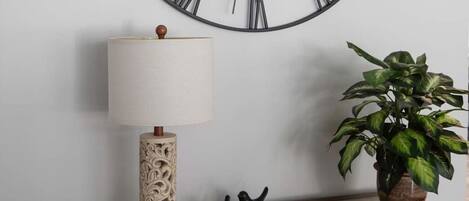 A wall-mounted clock with Roman numerals, a lamp, a potted plant, and a wooden side table with decorative items and books.