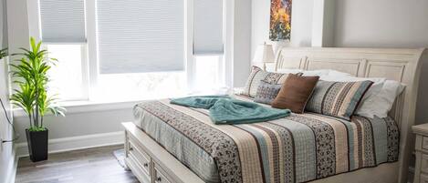 A bedroom with a bed, a nightstand, a potted plant, and a window with blinds.