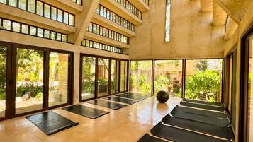 A spacious indoor area with multiple exercise mats arranged in rows, large windows allowing natural light, and a view of a lush garden.