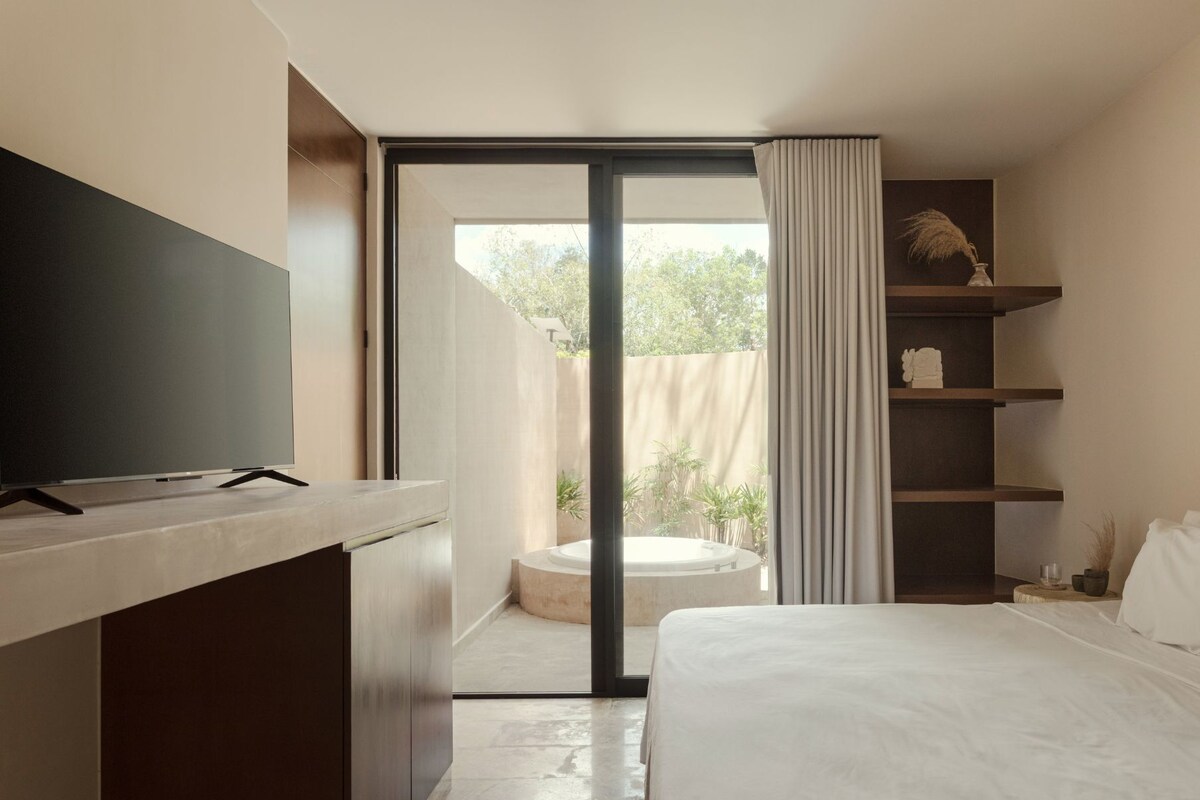 A modern hotel room with a large bed, a flat-screen TV, a small seating area with a round table, and a balcony with a view of greenery.