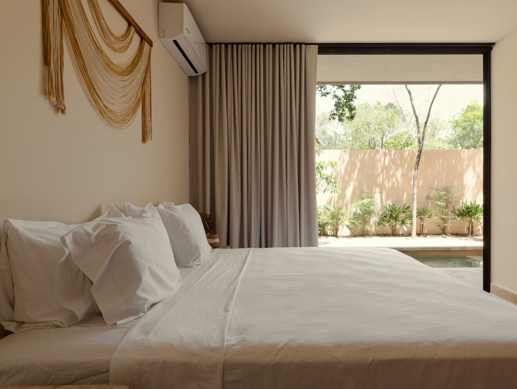 Quarto com cama, janela com cortinas e vista para área externa com plantas.