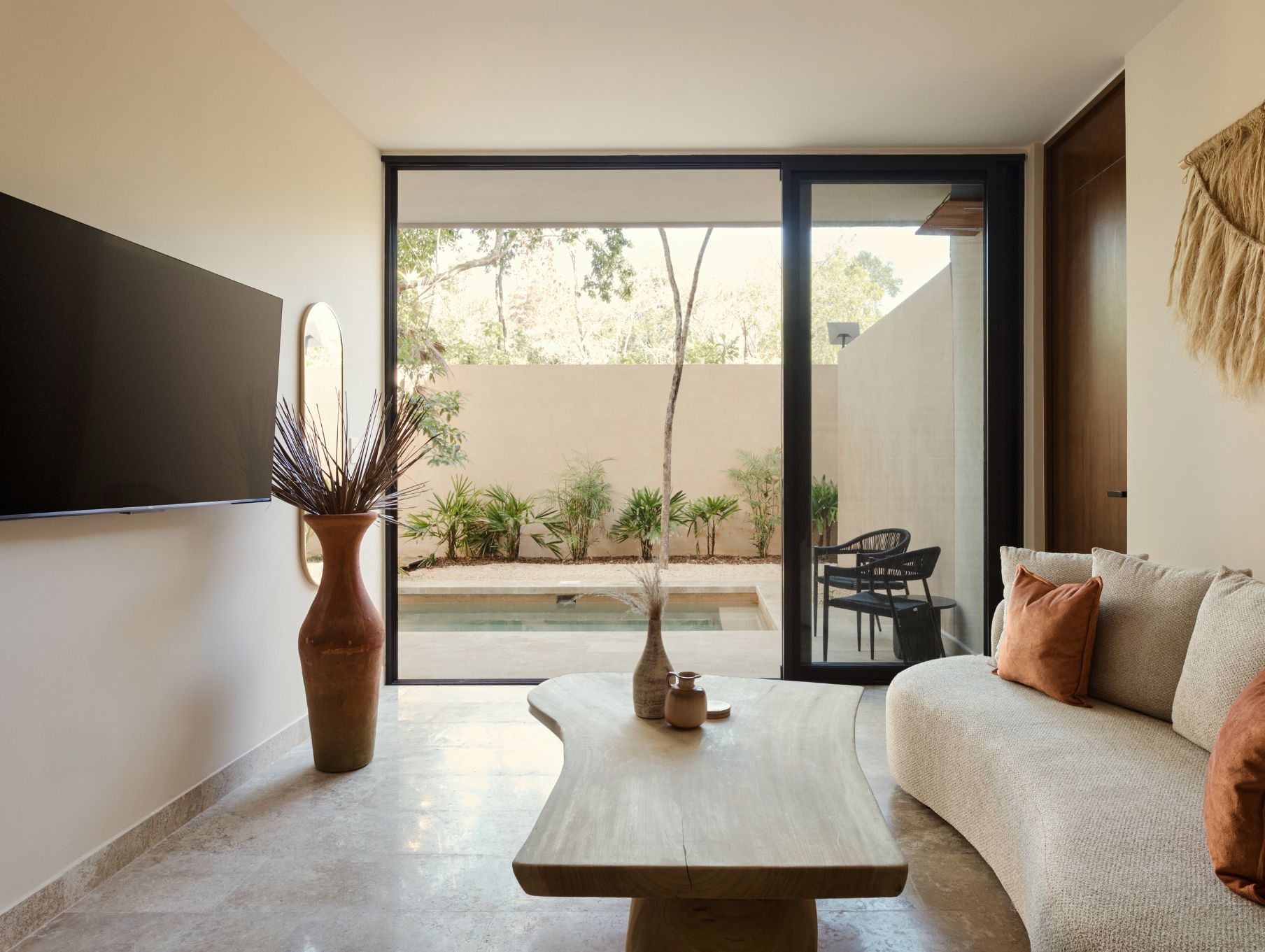 A modern living room with a sofa, a coffee table, and a large window offering a view of a pool and greenery.
