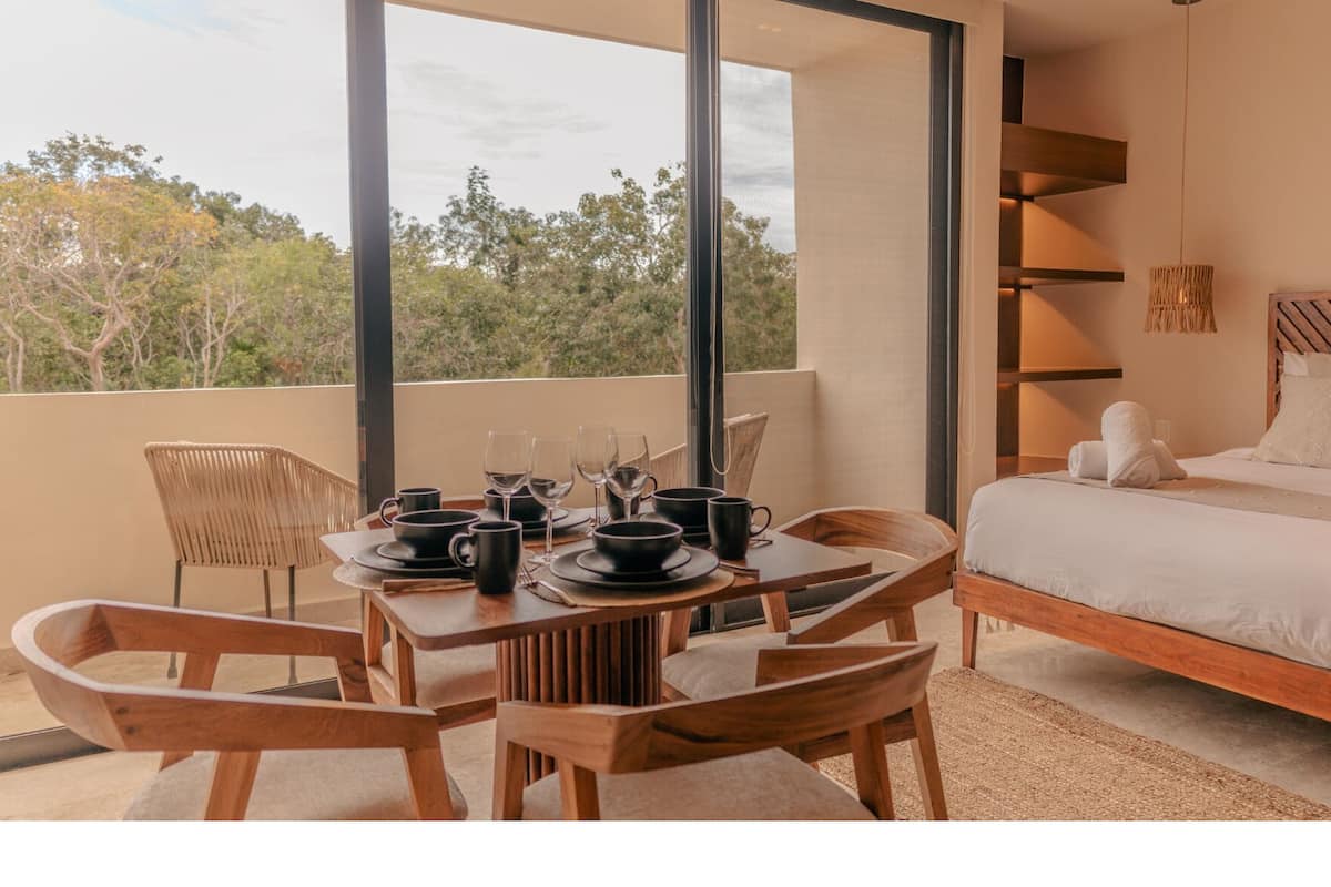 Una camera da letto moderna con un letto grande, una zona pranzo con un tavolo apparecchiato per due e vista sugli alberi attraverso ampie finestre.