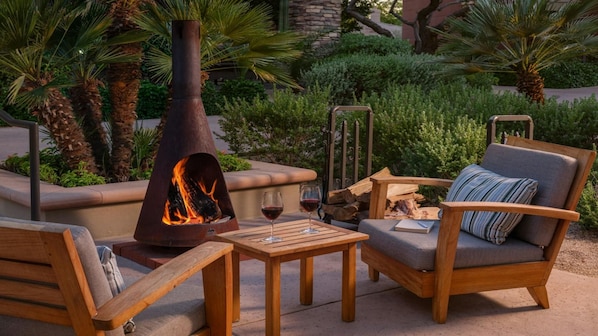 An outdoor seating area with a wood-burning stove, two glasses of red wine, and a wooden chair with a cushion.