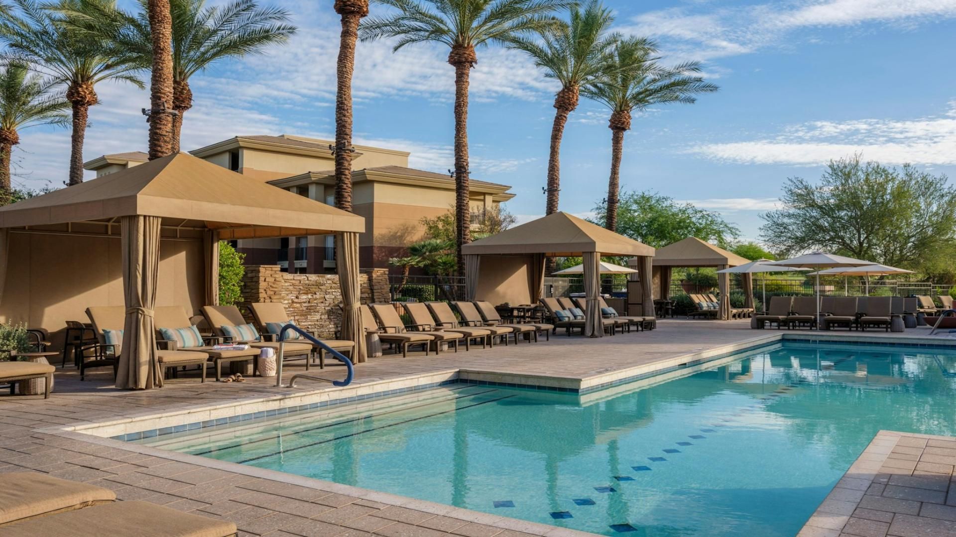A poolside area with lounge chairs and umbrellas, surrounded by palm trees and a building with a stone facade.