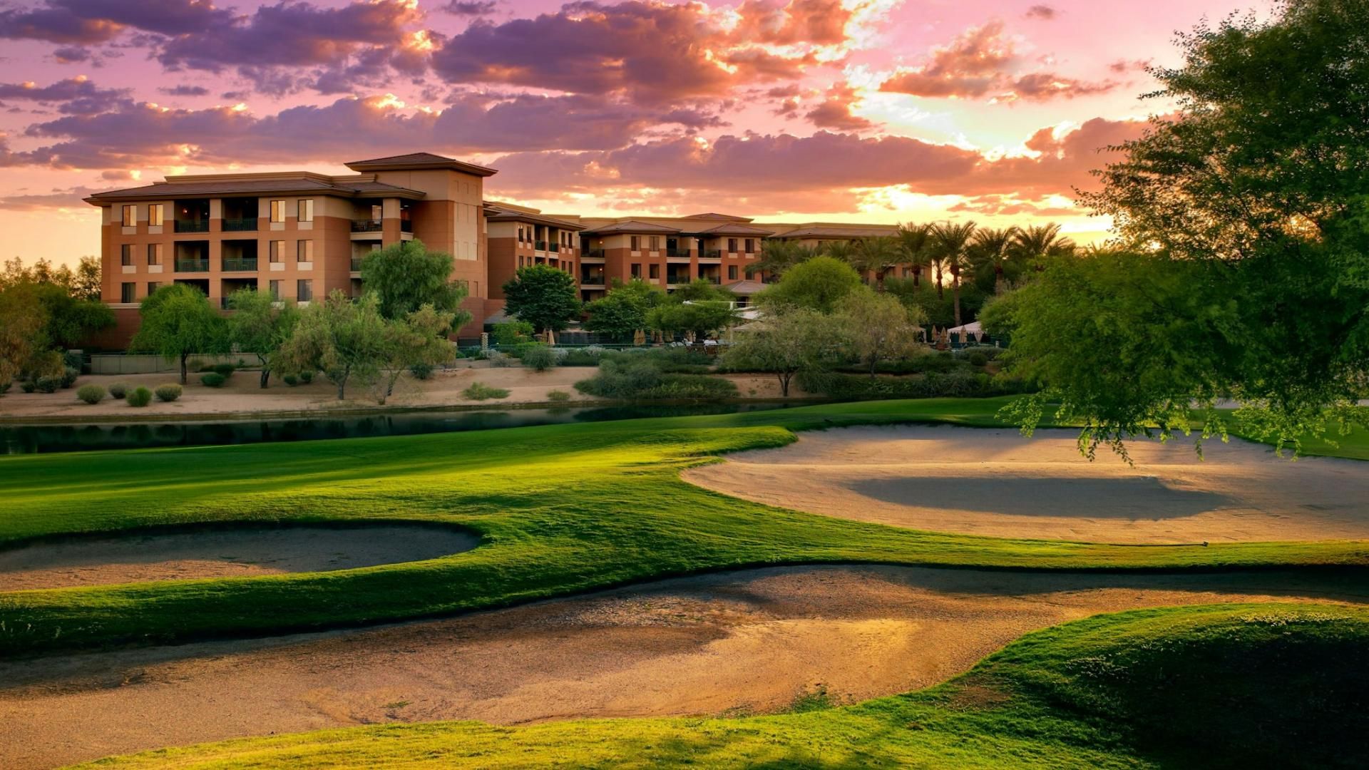 A golf course with bunkers and a building with multiple balconies in the background.