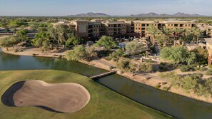Campo de golf con un cuerpo de agua, un puente y un complejo hotelero con varias construcciones.
