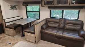 A compact RV interior with a dining area, a cushioned bench, a sofa, and a window offering a view of greenery. - Nature Lover's 2-Bedroom RV Escape (Hamilton)