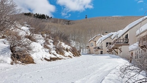 Una calle cubierta de nieve con edificios a la derecha, cielo despejado y una colina al fondo.