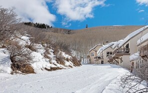Una strada innevata con edifici a destra, cielo sereno e una collina sullo sfondo.