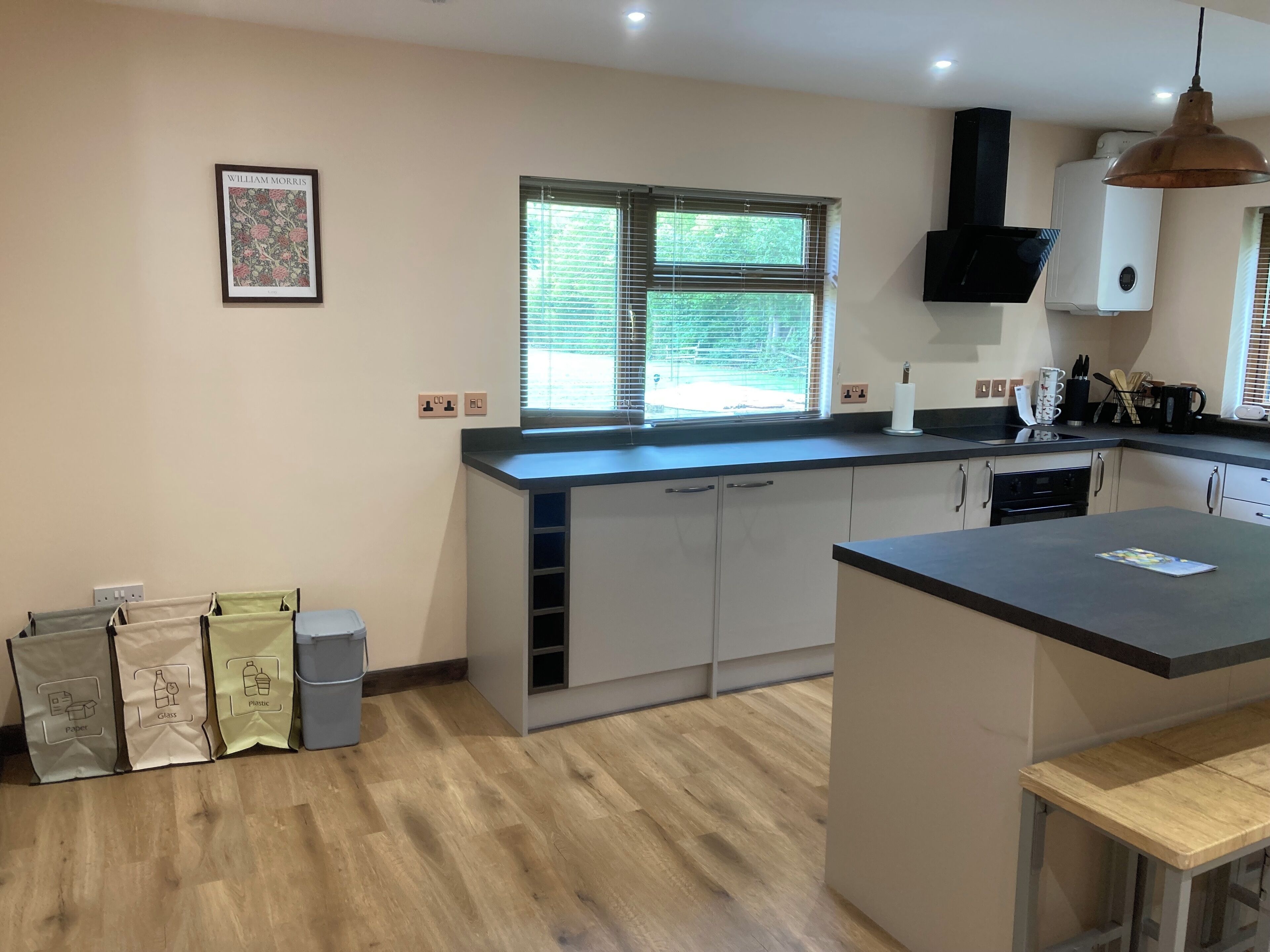 A modern kitchen with a central island, a built-in oven, and a window with blinds. There are paper, glass, and plastic recycling bags on the floor.