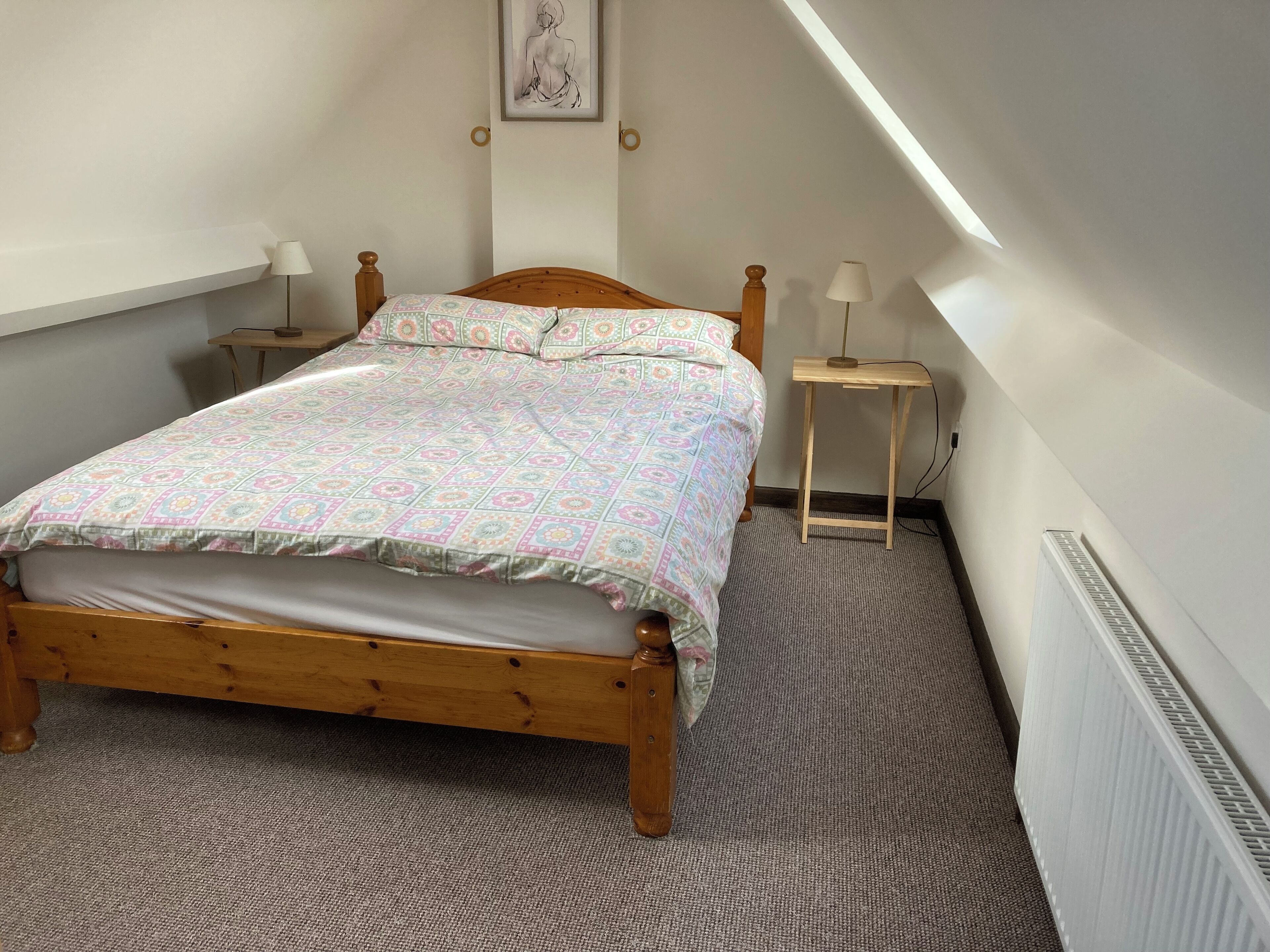 A bedroom with a wooden bed, patterned bedding, two bedside tables with lamps, a radiator, and a framed artwork on the wall.