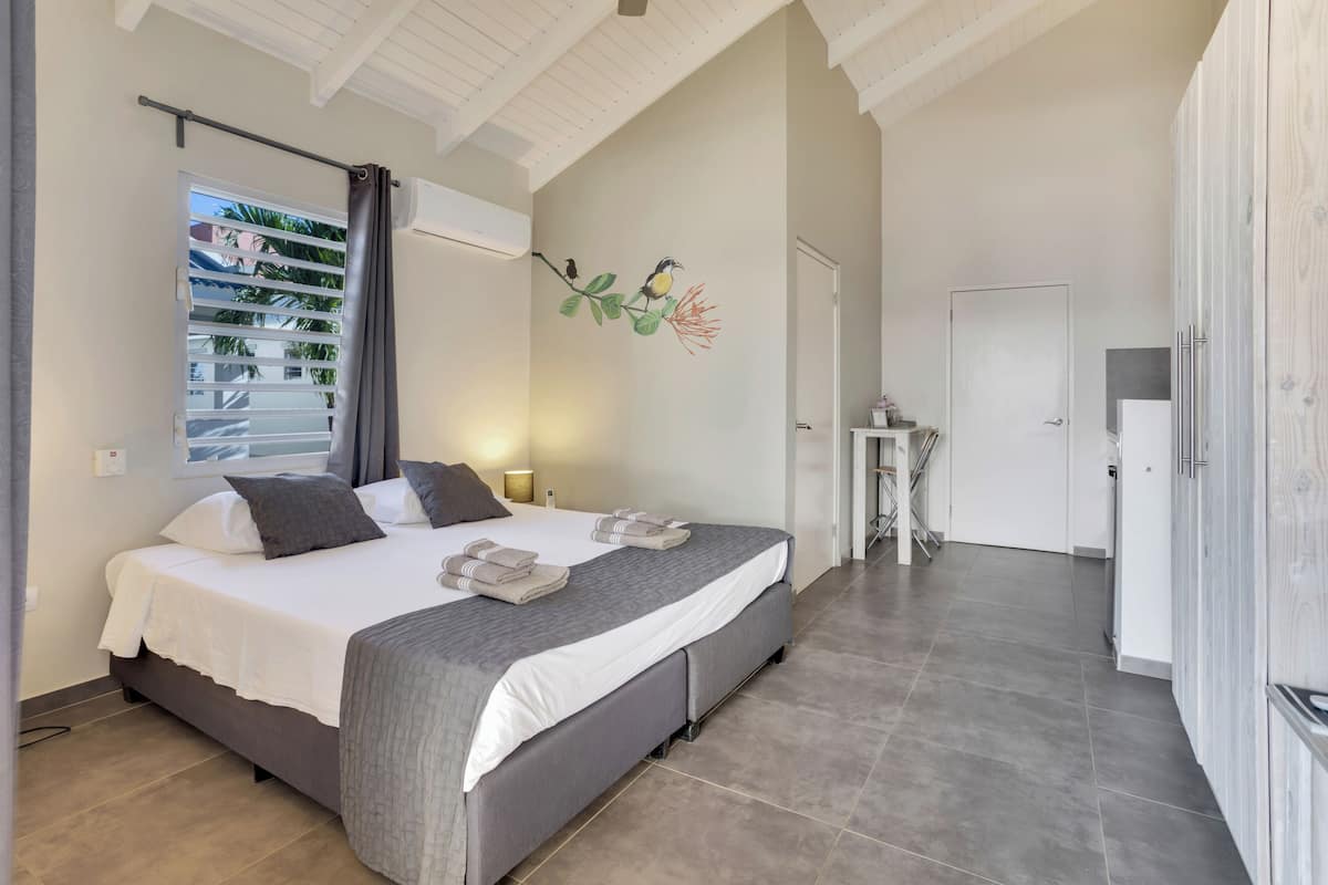 A bedroom with a large bed, a window with blinds, a wall-mounted air conditioner, and a small table with a lamp.