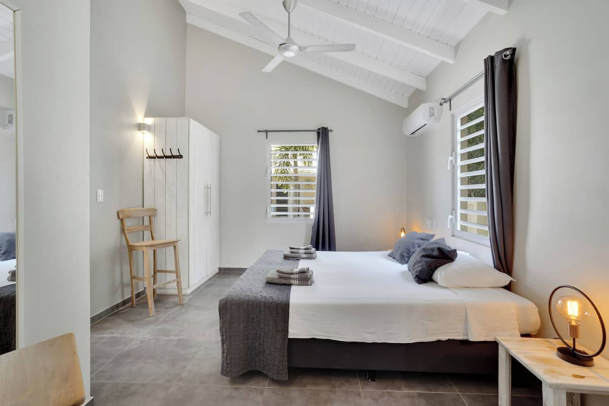 A bedroom with a bed, a chair, a bedside table with a lamp, a window with curtains, and an air conditioning unit.