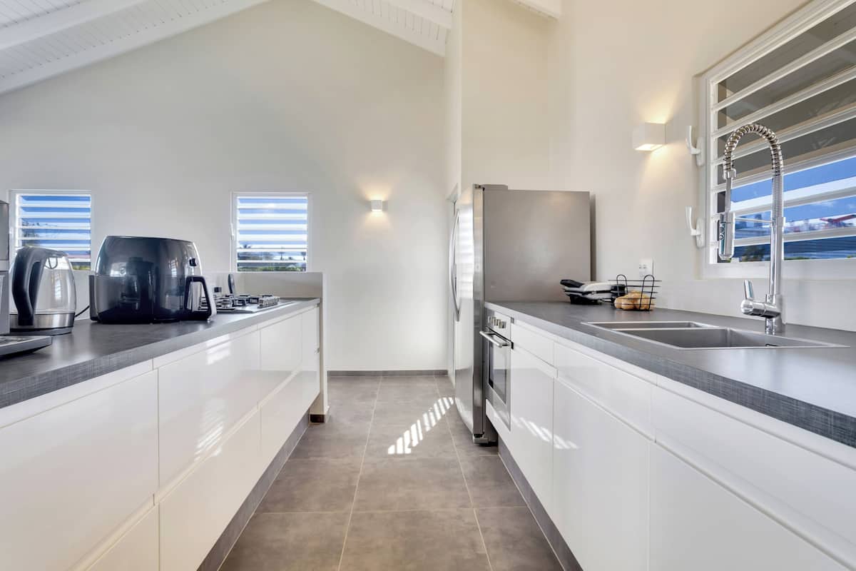 A modern kitchen with white cabinets, a stainless steel refrigerator, a built-in oven, and a large window providing natural light.