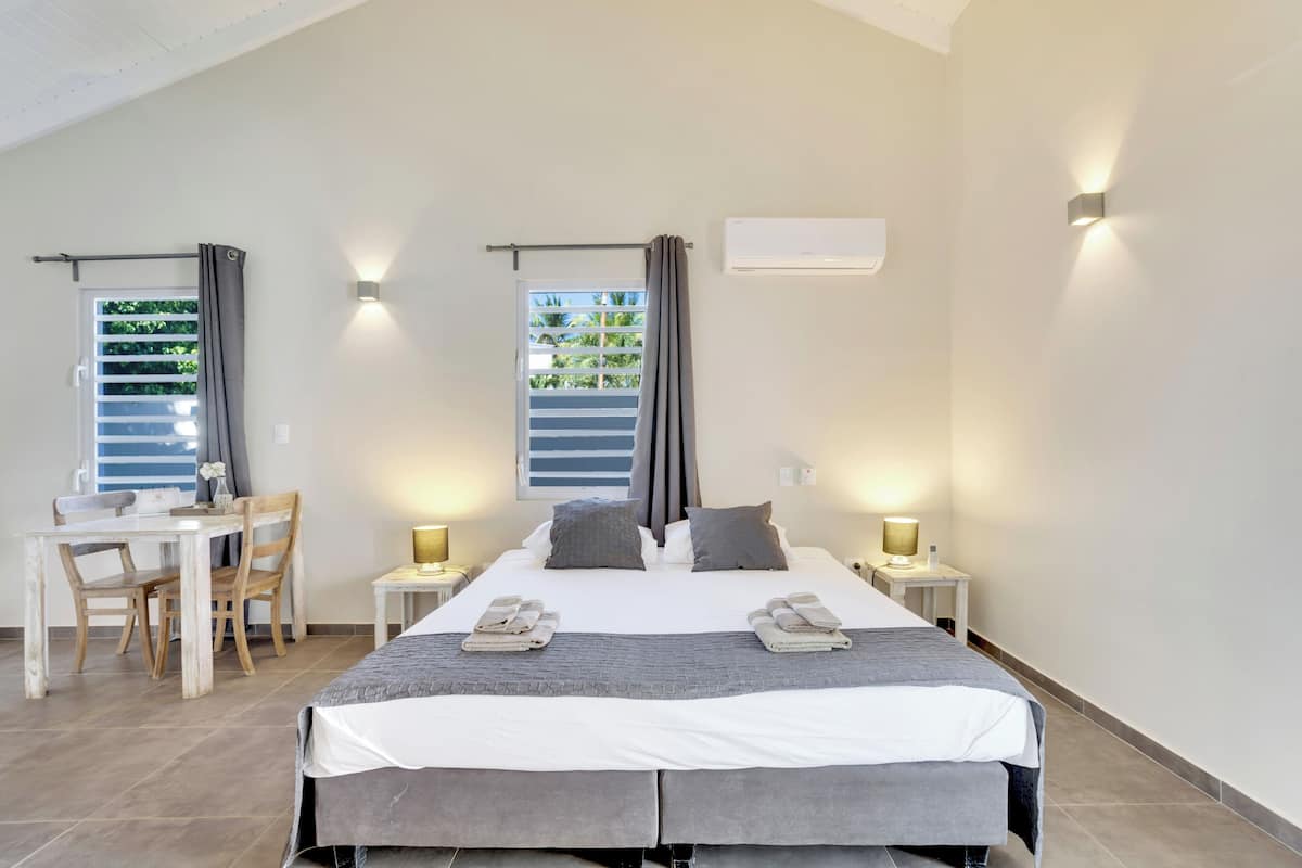 A hotel room with a bed, two bedside tables with lamps, a desk with chairs, and a window with curtains.